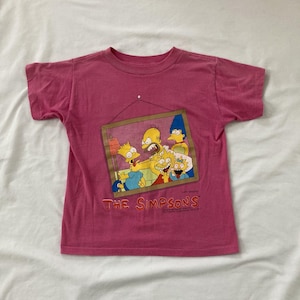 May include: A pink t-shirt featuring a framed image of The Simpsons family. The image shows Homer, Marge, Bart, Lisa, and Maggie Simpson in a cartoon style. The text "THE SIMPSONS" is printed in orange below the image.