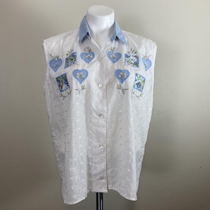 May include: White sleeveless button-down shirt with a light blue denim collar. The shirt features embroidered blue hearts and floral squares. The fabric has a delicate eyelet pattern throughout, adding texture and detail to the garment.
