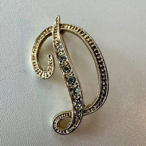 May include: A gold-toned brooch in the shape of the letter "D". The brooch is embellished with clear, sparkling rhinestones along the vertical stroke. The edges of the letter are detailed with a textured pattern.