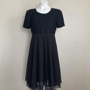May include: A black dress with a beaded bodice and short sleeves. The dress has a fitted waist and a flared skirt made of a flowing fabric. The dress is displayed on a mannequin against a neutral background.