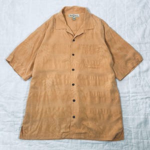 May include: A light orange button-down shirt with short sleeves. The shirt features a subtle palm tree pattern and a chest pocket. The shirt has a classic collar and is buttoned up with dark brown buttons. The shirt is made of a soft, textured fabric.