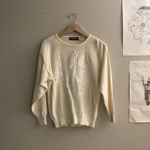 May include: Cream-colored sweater with a white bow and floral design. The long-sleeved crew neck sweater is hanging on a wooden hanger. The brand name "Dolcezza" is visible on the neckline.