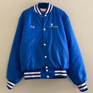 May include: A royal blue bomber jacket with white accents. The jacket features white stripes on the collar, cuffs, and hem. The name "Troy" is embroidered on the left chest, and the words "Mark Industries" are on the right chest.