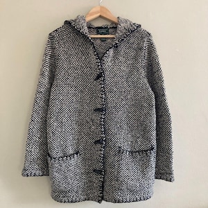 May include: A hooded, long-sleeved sweater in a black and white speckled pattern. The sweater has a button-up front with dark navy toggles, two front pockets, and navy trim. The sweater is hanging on a wooden hanger.
