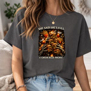 May include: Dark gray t-shirt with a graphic of a stack of burgers, fries, and drinks, with skeleton hands reaching up. The text on the shirt reads "SHE SAID SHE'S FULL" and "I ORDERED MORE".
