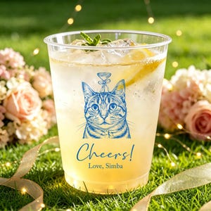 May include: A clear plastic cup filled with a light yellow beverage, ice cubes, and a lemon slice. The cup features a blue line-art illustration of a cat with a small martini glass above its head, with the text Cheers! Love, Simba below.