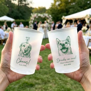 May include: Two frosted plastic cups featuring green ink illustrations of dogs. The left cup shows a dog with a glass on its head and the text "Cheers! Love, Bella". The right cup shows a dog and the text "Drinks on me! Cheers to Mom & Dad! 09.23.2026".