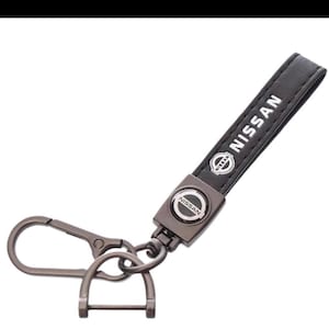 May include: Black leather Nissan keychain with a metal clasp and a metal plate with the Nissan logo. The keychain strap has the word "NISSAN" printed in white.