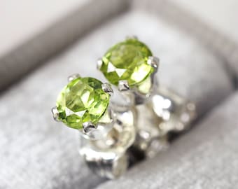 May include: A pair of stud earrings featuring round, faceted, lime green peridot gemstones set in silver-toned metal prongs. Each earring has a secure metal backing and is displayed in a soft, grey, velvet-lined jewelry box container.