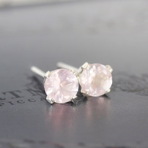 Peut inclure: Une paire de boucles d'oreilles à clous avec des pierres précieuses en quartz rose rondes et facettées, montées sur des griffes en métal argenté. Les pierres sont translucides avec un léger éclat, et les tiges argentées sont visibles à l'arrière.