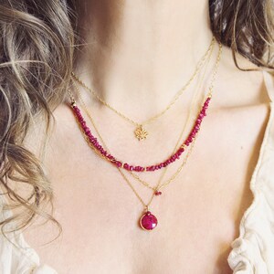 May include: A multi-layered necklace set with gold chains and ruby-coloured gemstone beads. The necklaces include a lotus flower pendant and a round ruby pendant, creating a delicate and elegant jewellery ensemble.