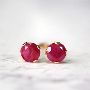 May include: A pair of ruby stud earrings. Each earring features a faceted, round, deep red gemstone set in a gold-colored bezel. The earrings are displayed on a textured, off-white surface.
