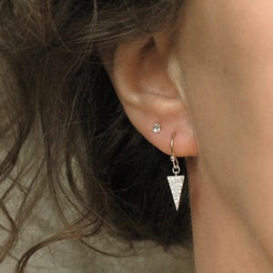 May include: A silver-toned earring featuring a small, downward-pointing triangular pendant encrusted with tiny clear stones. The earring hangs from a thin hook wire, and a small, round, clear stone stud is visible in the upper earlobe area.