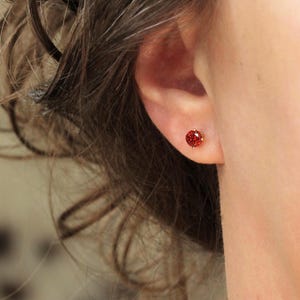 May include: A close-up of an ear with a small, round, red gemstone stud earring. The earring is set in a gold-coloured metal. The person's brown hair is slightly out of focus in the background.