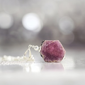 May include: A silver chain necklace with a raw pink ruby pendant. The ruby is a natural, rough-cut gemstone with a deep pink colour.