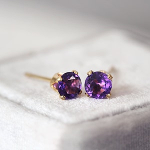 Könnte beinhalten: Ein Paar Ohrstecker mit runden, tiefvioletten Amethyst-Edelsteinen, gefasst in goldfarbenem Metall. Die Ohrringe werden auf einer weichen, weißen Oberfläche präsentiert, die ihre lebendige Farbe und ihr Design hervorhebt.