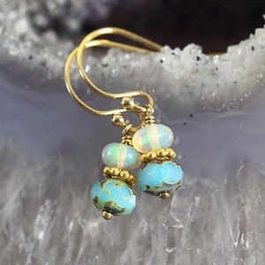 May include: Gold-coloured earrings with a delicate design. Each earring features three beads: an opal bead, a middle bead, and a blue and green patterned bead. The earrings have a hook closure.