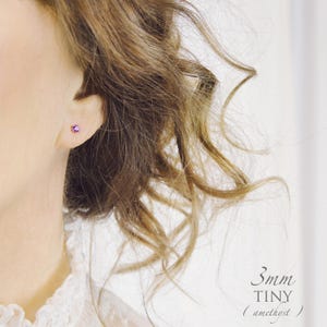 May include: A small stud earring featuring a round, purple amethyst gemstone. The text 3mm TINY (amethyst) is visible in the lower right corner. The earring is worn in an earlobe, with the gemstone centred against the skin.