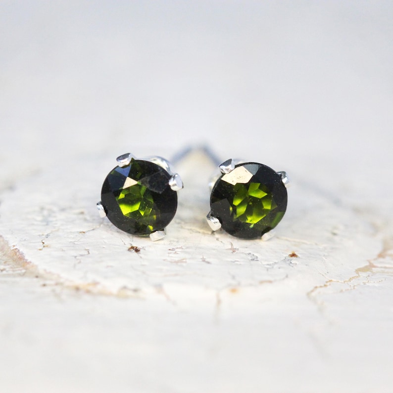 May include: A pair of stud earrings with round, faceted, olive-green gemstones set in silver-coloured metal. The earrings are displayed on a textured, off-white surface, highlighting their colour and design.
