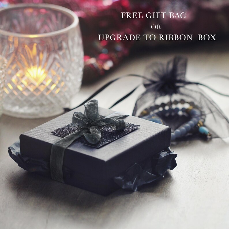 Puede incluir: Una caja de regalo negra con una parte superior brillante y una cinta verde oscuro. La caja est&aacute; sobre papel de seda negro. Un portavelas de cristal transparente y una bolsa de malla negra con joyas est&aacute;n en el fondo.
