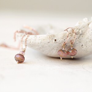 May include: A rose gold necklace and matching earrings set. The necklace features a pendant with a large faceted bead and smaller beads. The earrings have a similar design with faceted beads and rose gold hooks. The jewelry is displayed on a white, textured surface.