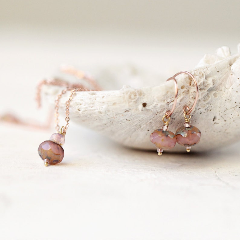 May include: A rose gold necklace and matching earrings set. The necklace features a pendant with a large faceted bead and smaller beads. The earrings have a similar design with faceted beads and rose gold hooks. The jewelry is displayed on a white, textured surface.