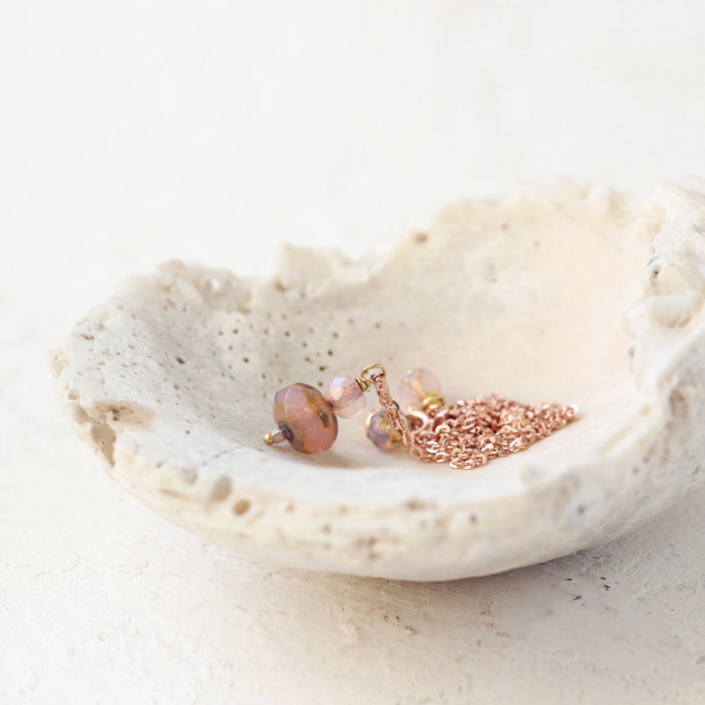 May include: A delicate necklace with a rose gold chain and pendant, featuring faceted pink and peach-colored beads. The jewelry rests inside a white, textured seashell, creating a soft, elegant display. The necklace is a perfect accessory for any occasion.