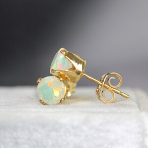 Puede incluir: Un par de pendientes de oro con piedras preciosas de &oacute;palo iridiscente. Los pendientes tienen un dise&ntilde;o cl&aacute;sico de poste y cierre. Los &oacute;palos muestran una gama de colores, incluyendo blanco, amarillo y verde. Los pendientes est&aacute;n sobre un fondo blanco suave.