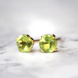 May include: A pair of stud earrings featuring round, light green peridot gemstones set in gold-colored prongs. The earrings are displayed on a light, textured surface, showcasing their vibrant color and simple design.