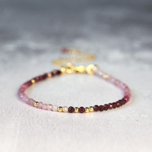 May include: A delicate bracelet featuring a gradient of faceted gemstone beads transitioning from deep burgundy to pale pink, interspersed with small, polished gold-tone metal beads. The piece includes a gold-tone metal chain and a lobster clasp.
