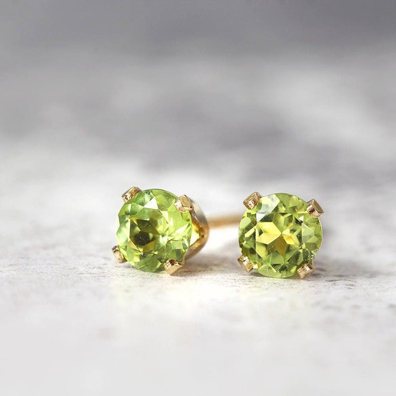 A pair of stud earrings featuring faceted, sparkling, vibrant green peridot gemstones set into a gold prong setting. The earrings are round in shape, with ear post fitting and butterfly backs. The earrings are simple and minimal in style.