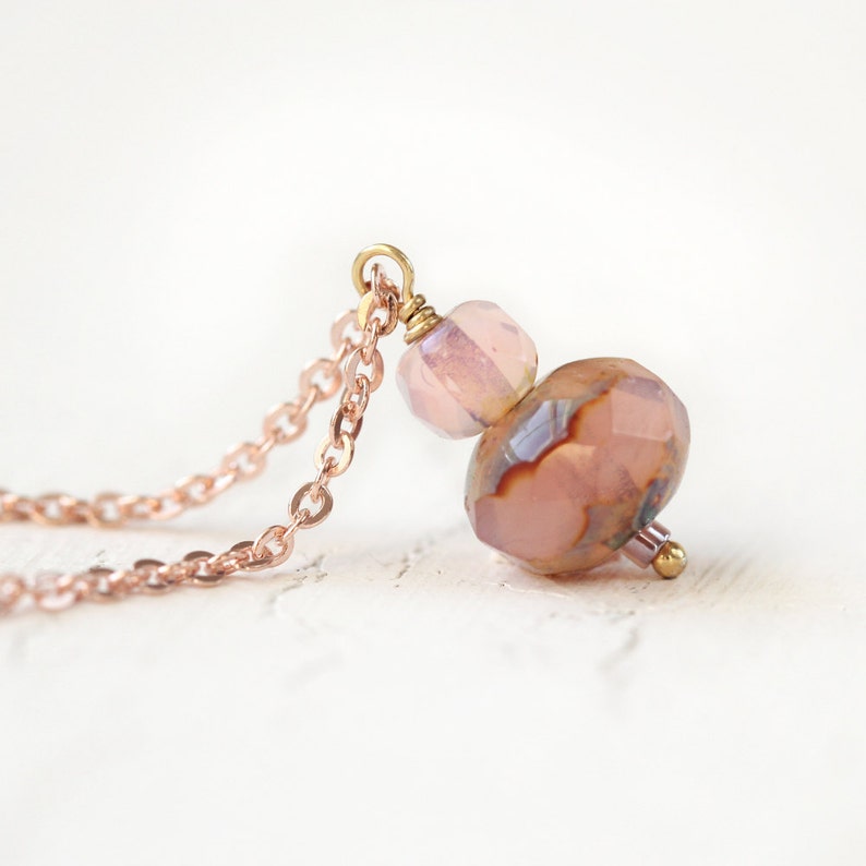 May include: A delicate necklace featuring a rose gold chain and pendant. The pendant is composed of faceted, light pink and brown beads with gold accents. The necklace is displayed against a white background.
