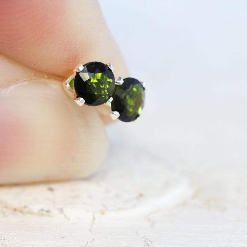 May include: A pair of oval-cut, dark green gemstone stud earrings. The gemstones are set in a silver-coloured metal with four prongs. The earrings are displayed against a soft, blurred background.