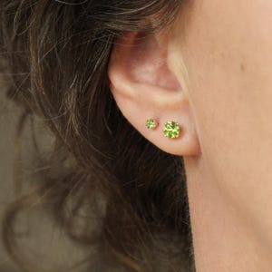 May include: Two round, faceted, lime green peridot stud earrings set in gold metal prongs. The larger earring is positioned on the lower earlobe, while the smaller earring is placed slightly higher on the ear, both featuring a brilliant cut.