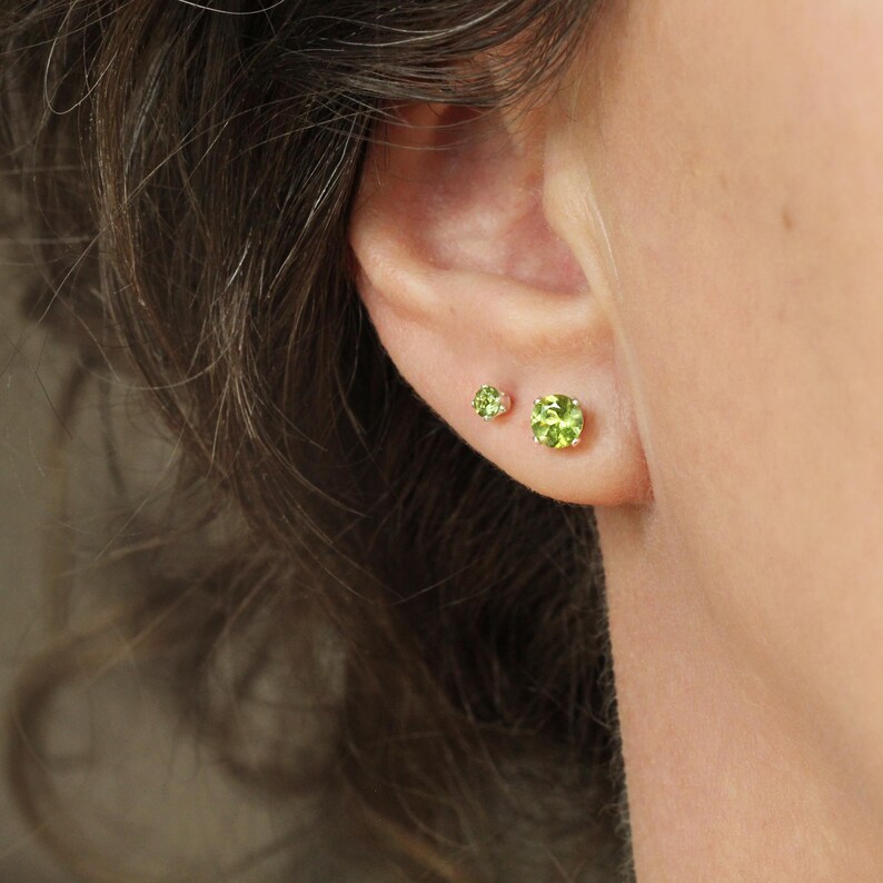 May include: Two round, faceted, lime green peridot stud earrings set in gold metal prongs. The larger earring is positioned on the lower earlobe, while the smaller earring is placed slightly higher on the ear, both featuring a brilliant cut.