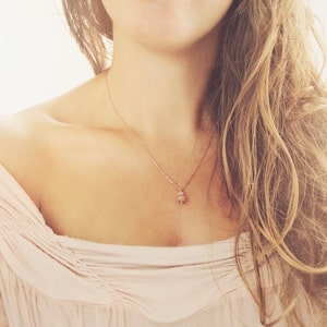 May include: A delicate rose gold necklace with a small, light pink pendant. The necklace is worn on a person with fair skin, wearing a light pink off-the-shoulder top. The chain is fine and the pendant is a small, rounded shape.