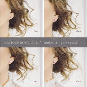 May include: A stud earring size guide from Artique Boutique. The image shows four different sizes of stud earrings: 3mm, 4mm, 5mm, and 6mm. Each earring is a small, round, purple gemstone. The earrings are shown on a person's ear.
