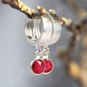 May include: A pair of silver hoop earrings with dangling red gemstone charms. The earrings are hanging from a branch.