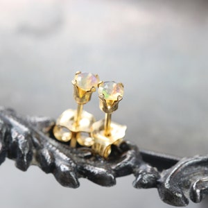 Puede incluir: Un par de pendientes de oro con piedras preciosas de &oacute;palo iridiscente. Los pendientes est&aacute;n engastados en un engaste cl&aacute;sico de cuatro puntas. Los pendientes se exhiben sobre una hoja de metal decorativa de color gris oscuro.