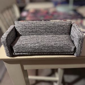 Puede incluir: Un sofá en miniatura de tweed gris y blanco con un cojín trasero y reposabrazos a juego. El pequeño sofá está colocado sobre una mesa de madera de color claro. El fondo está borroso, lo que sugiere un entorno interior.