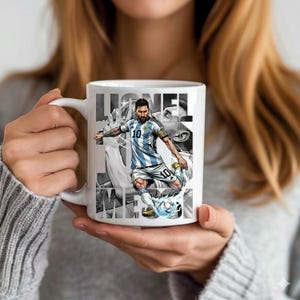 May include: White ceramic mug featuring a graphic of a soccer player in a blue and white striped jersey, kicking a soccer ball. The mug has the name "Lionel Messi" printed in a stylized font.
