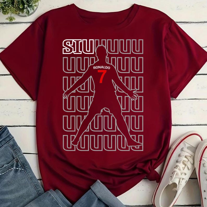 May include: Maroon t-shirt featuring a silhouette of a soccer player with the name "Ronaldo" and the number "7" in red. The word "SIUUUU" is printed in white above the silhouette. White sneakers are visible in the bottom right corner.