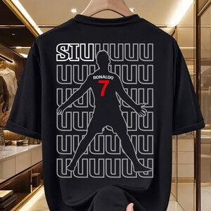 May include: Black t-shirt featuring a silhouette of a soccer player with the name "Ronaldo" and the number "7" in red. The word "SIU" is at the top, with a repeating pattern of "UUUU" in the background. The shirt is hanging on a wooden hanger.