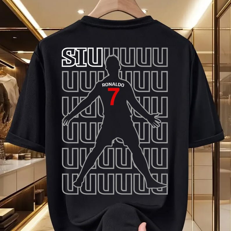 May include: Black t-shirt featuring a silhouette of a soccer player with the name "Ronaldo" and the number "7" in red. The word "SIU" is at the top, with a repeating pattern of "UUUU" in the background. The shirt is hanging on a wooden hanger.