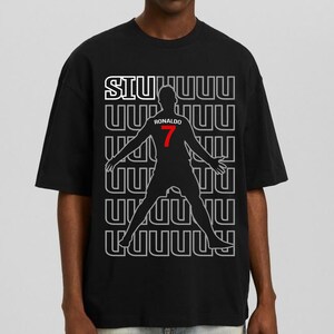 May include: Black t-shirt featuring a silhouette of a soccer player with the number 7 in red. The shirt has the word "SIU" in white and the player's name "Ronaldo" above the number. The background has a repeating pattern.