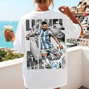 May include: White t-shirt featuring a graphic of a soccer player in a blue and white striped jersey, kicking a soccer ball. The player's name, "Lionel Messi," is printed in large, bold letters above and below the image.