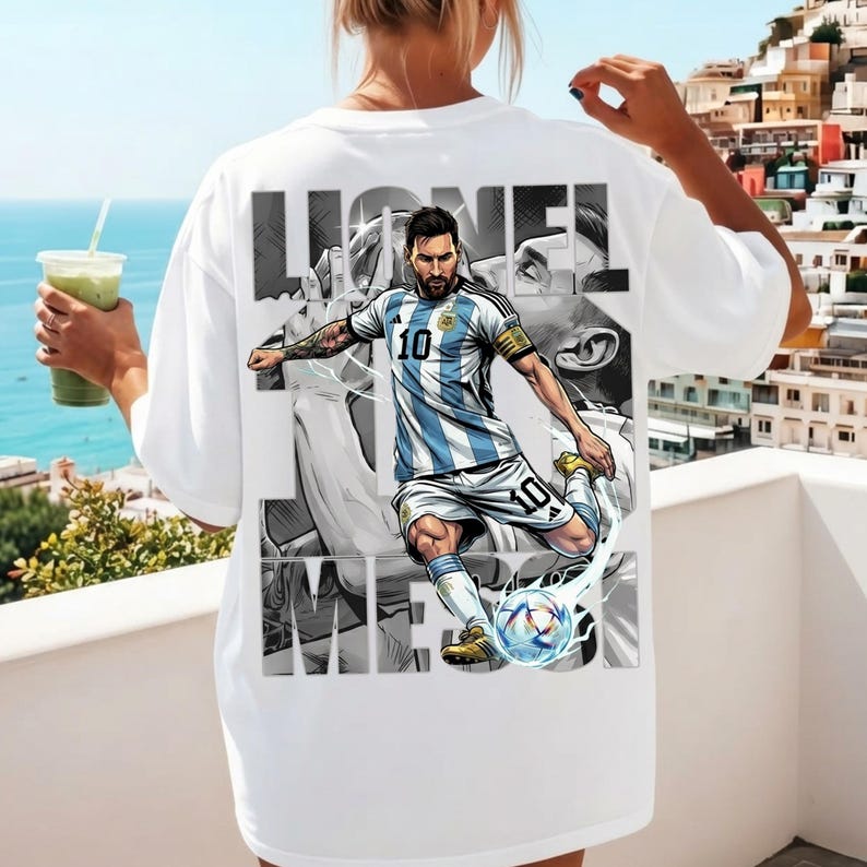 May include: White t-shirt featuring a graphic of a soccer player in a blue and white striped jersey, kicking a soccer ball. The player's name, "Lionel Messi," is printed in large, bold letters above and below the image.