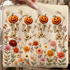 May include: A beige sweatshirt featuring a graphic print of four skeletons with jack-o'-lantern heads dancing among various colorful flowers including red, orange, and pink blossoms with green stems and leaves on a light cream background.