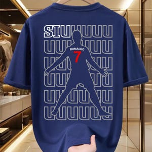 May include: Navy blue t-shirt featuring a silhouette of a soccer player with the name "RONALDO" and the number "7" in red. The word "SIU" is printed in white above the player. The design is repeated in a pattern.