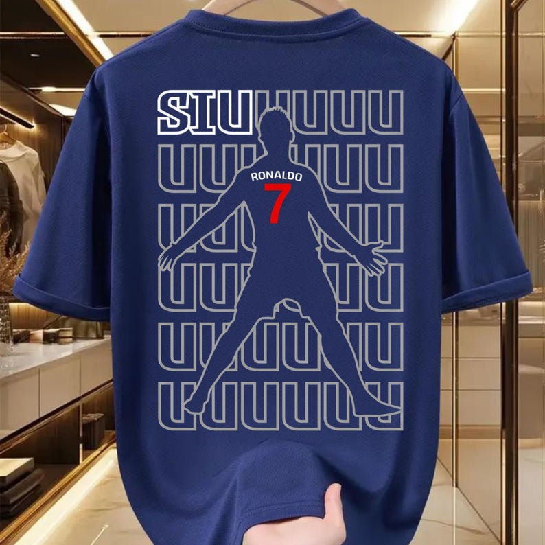 May include: Navy blue t-shirt featuring a silhouette of a soccer player with the name "RONALDO" and the number "7" in red. The word "SIU" is printed in white above the player. The design is repeated in a pattern.